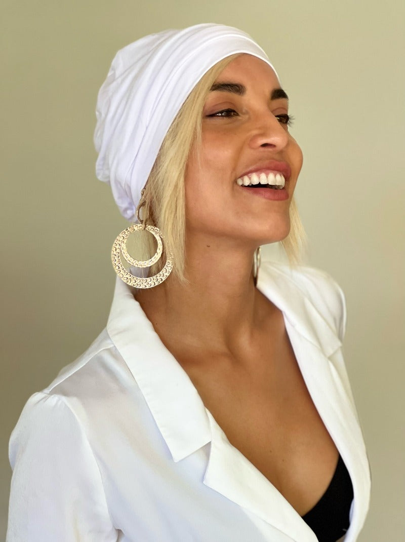 Woman wearing creamy blonde human hair hat wig under a white softie beanie, with a white shirt and gold earrings